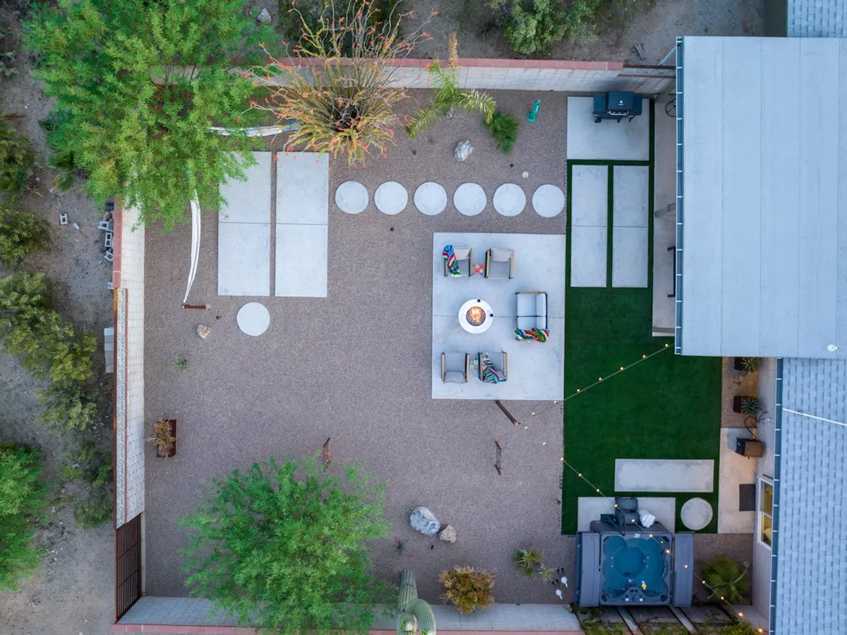 An inviting outdoor scene featuring a circular fire pit surrounded by modern seating, a hot tub nestled nearby, and a stone pathway leading through a landscaped yard. String lights provide a warm glow, suggesting a welcoming and intimate space for evening gatherings or quiet relaxation under the stars.