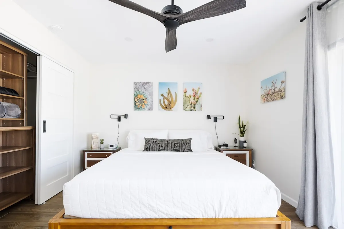 A serene bedroom with a comfortable bed adorned with soft linens, tasteful artwork above the headboard, nightstands on either side, and a ceiling fan overhead. Natural light filters gently into the space, creating a tranquil atmosphere ideal for relaxation and rejuvenation.