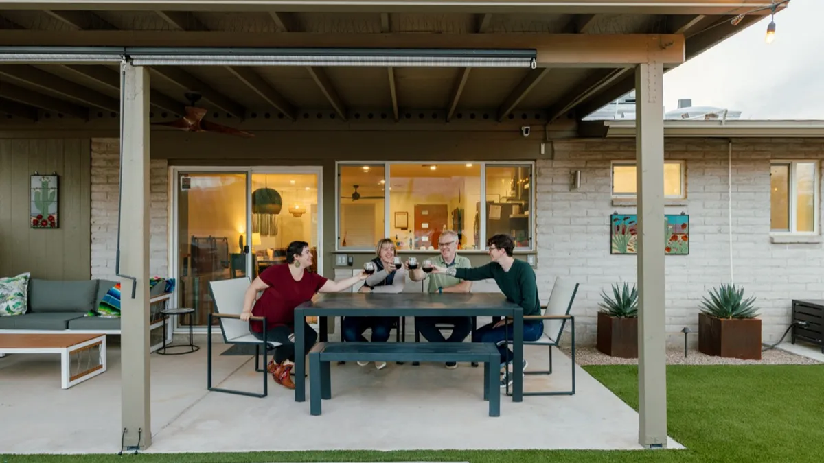 This image shows a lively group of friends gathered around an outdoor dining table on a green lawn. Warm interior lighting glows from the house, and there is a cheerful atmosphere as guests enjoy dining and socializing together in the fresh air.