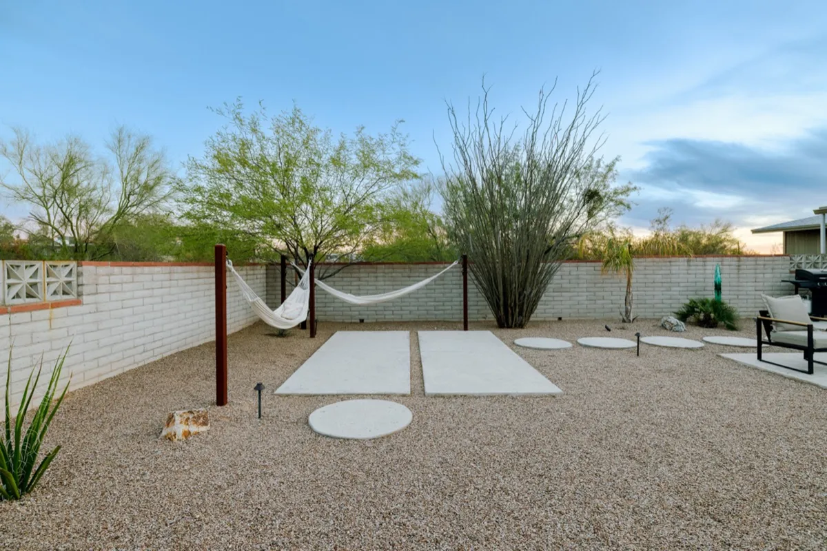 A serene outdoor space with a hammock strung between posts, surrounded by decorative desert landscaping and paved walkways. The area feels secluded and peaceful, inviting relaxation, meditation, or a quiet moment shared between two people while watching the sunset.