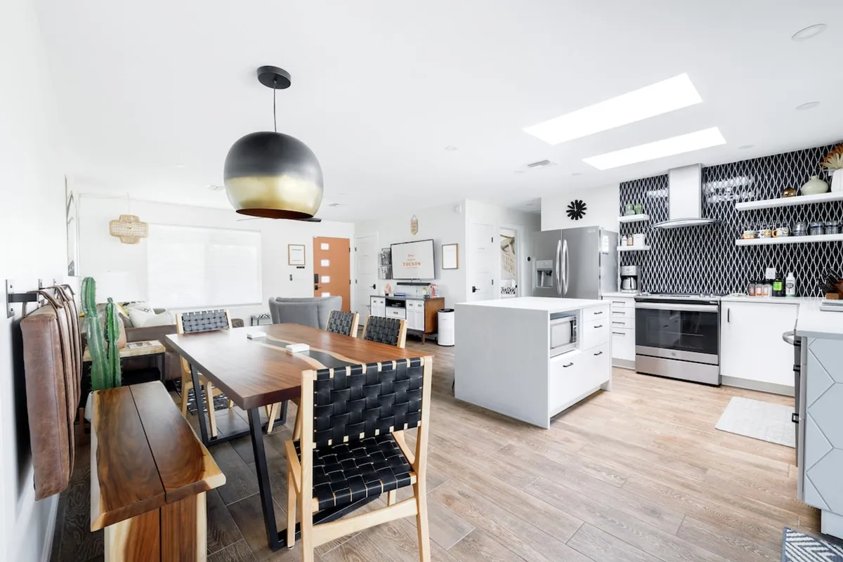 A large wooden dining table set for a meal stands adjacent to a sleek kitchen island. The black and white patterned backsplash adds a modern touch, and a stylish hanging light fixture creates a warm, inviting glow. The scene suggests room for both casual gatherings and celebratory dinners, with seamless flow between dining and kitchen spaces.