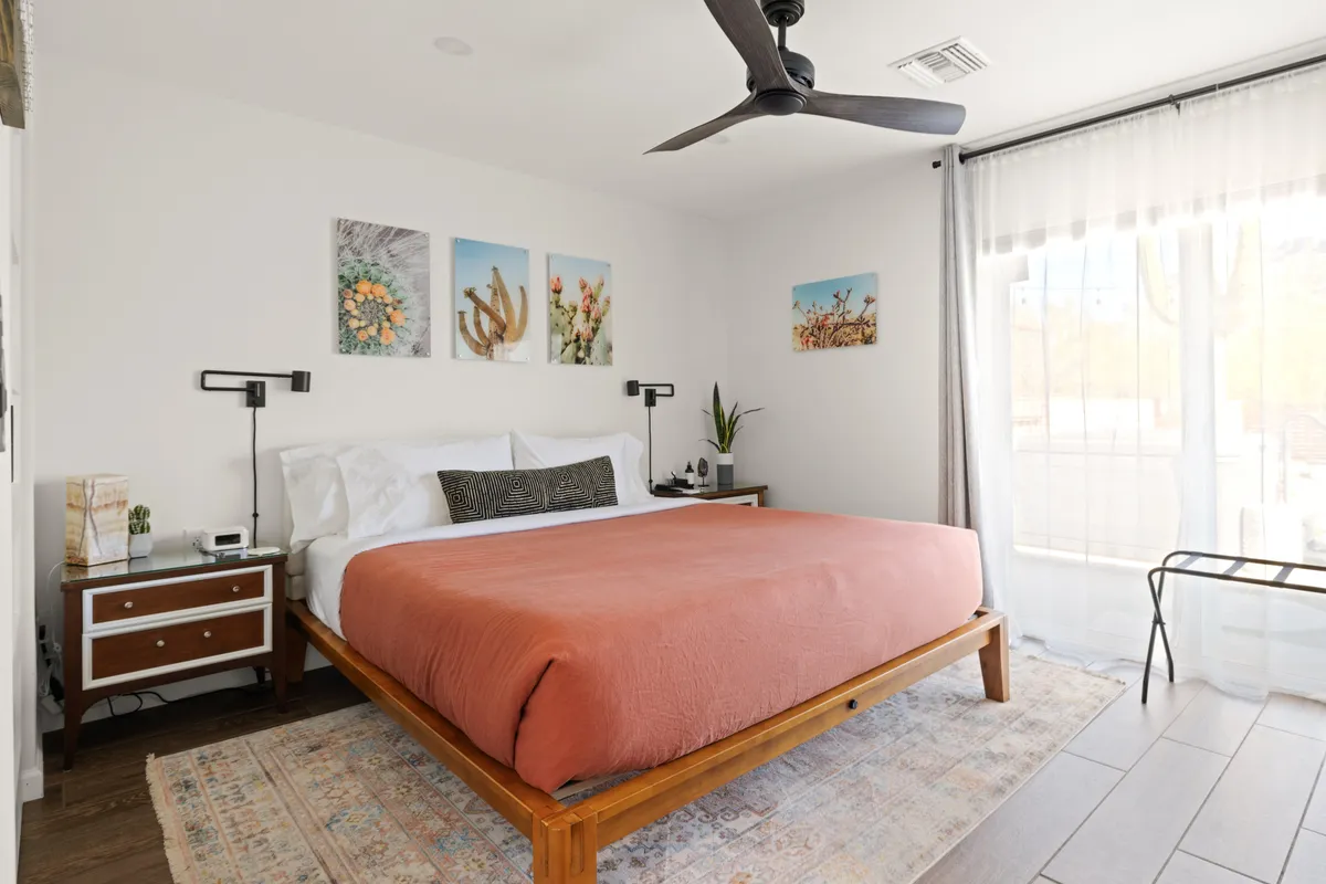 A cozy bedroom showcasing a bed with an orange coverlet, vibrant artwork on the wall, and abundant natural light streaming through the window. The room feels intimate and infused with personality, echoing local Tucson color palettes.