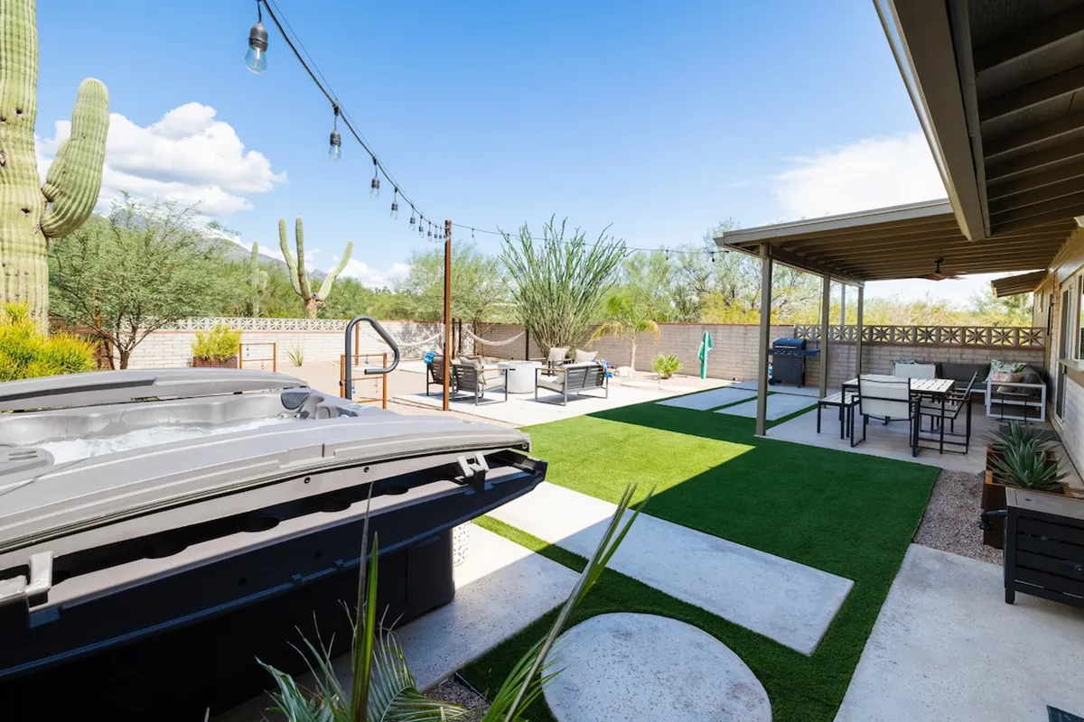 A tranquil backyard scene featuring a bubbling hot tub, lush lawn, inviting seating arrangements, decorative string lights, and cacti. The setting sun casts a warm glow, inviting guests to relax and enjoy the outdoor amenities, with clear views of the surrounding landscape.