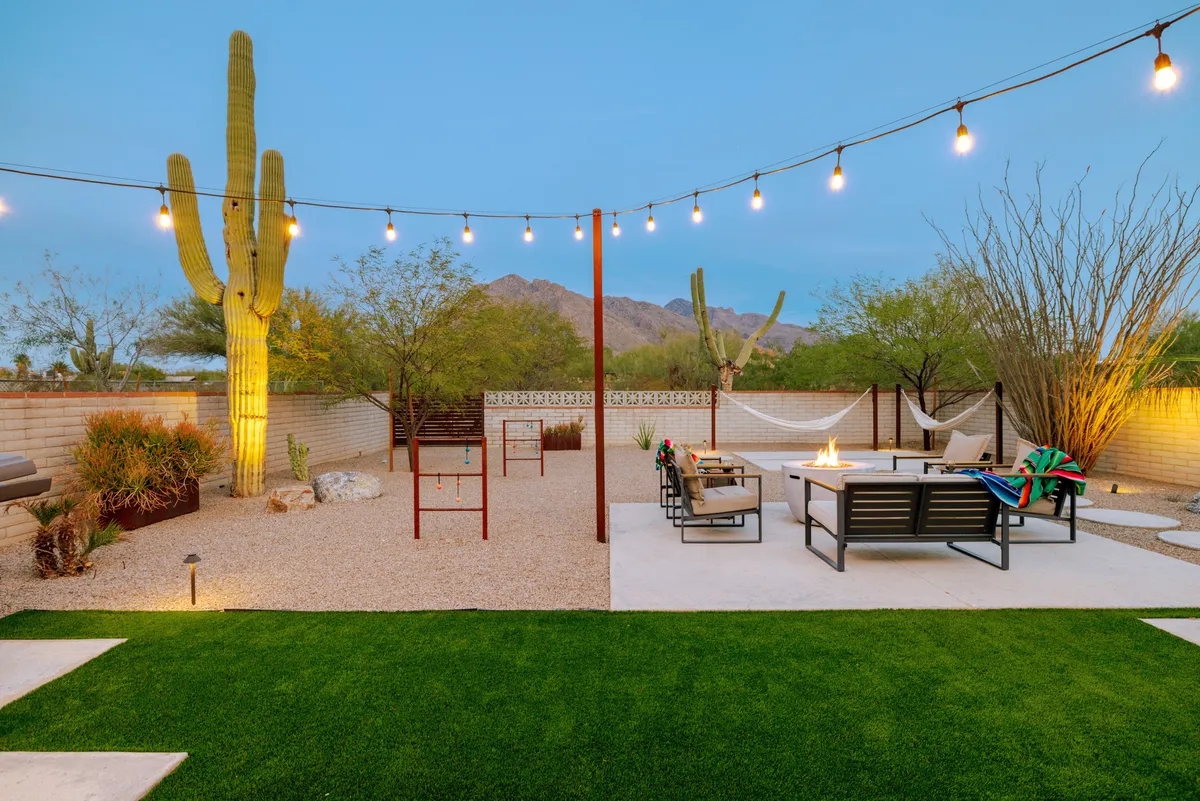 The image showcases a tranquil desert backyard oasis, framed by lush cacti and twinkling string lights. Central to the scene is a cozy fire pit surrounded by inviting outdoor seating, with a hammock gently swaying nearby—all set against the backdrop of Tucson’s breathtaking mountain vistas.