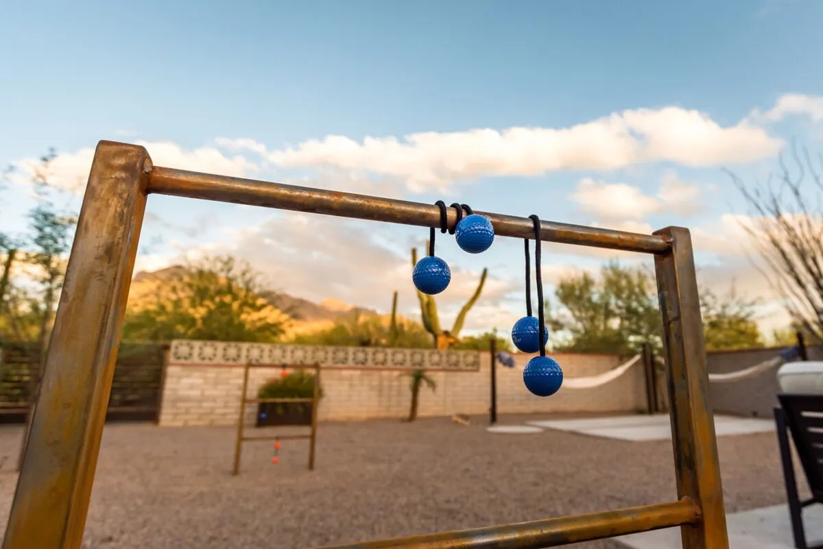 A playful and inviting backyard view featuring a rack of blue balls set up for an outdoor game, a hammock gently swaying nearby, and expansive mountain vistas in the background. Cacti and desert landscaping further contribute to the sense of a true desert oasis.