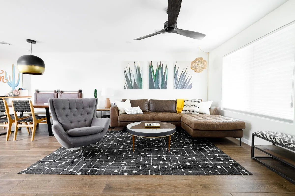 A spacious, open living area featuring a stylish brown sectional sofa, a gray armchair, a round coffee table, textured rug, and striking wall art. The room is illuminated by natural light, creating a warm, inviting atmosphere ideal for relaxation and conversation.