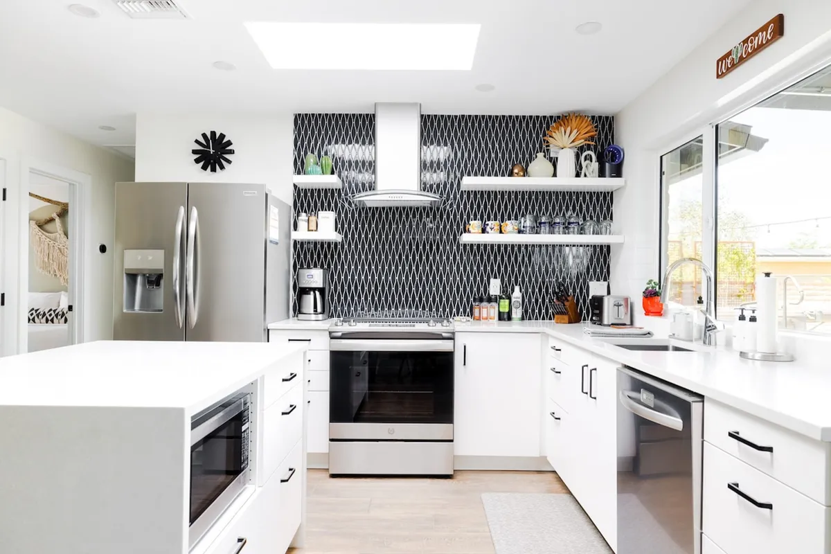 A modern kitchen features stainless steel appliances, geometric backsplash, open shelving, and a skylight that bathes the space in natural light. The kitchen island provides ample prep space and serves as a natural gathering point for entertaining or meal preparation.