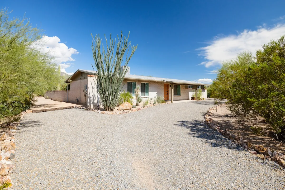 The exterior of the house, featuring a welcoming main entrance framed by tall cacti and a gravel driveway, all set against a tranquil desert backdrop. The porch is positioned to take in the sunset and enjoys the ambiance of the surrounding landscape, inviting guests to explore or relax.