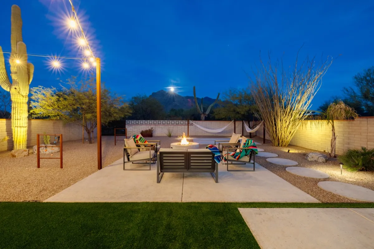 A cozy outdoor scene with a fire pit encircled by lounge seating and hammocks, all set under string lights. The foreground features a cactus and comfortable chairs, perfect for relaxing, storytelling, or stargazing in the desert evening.